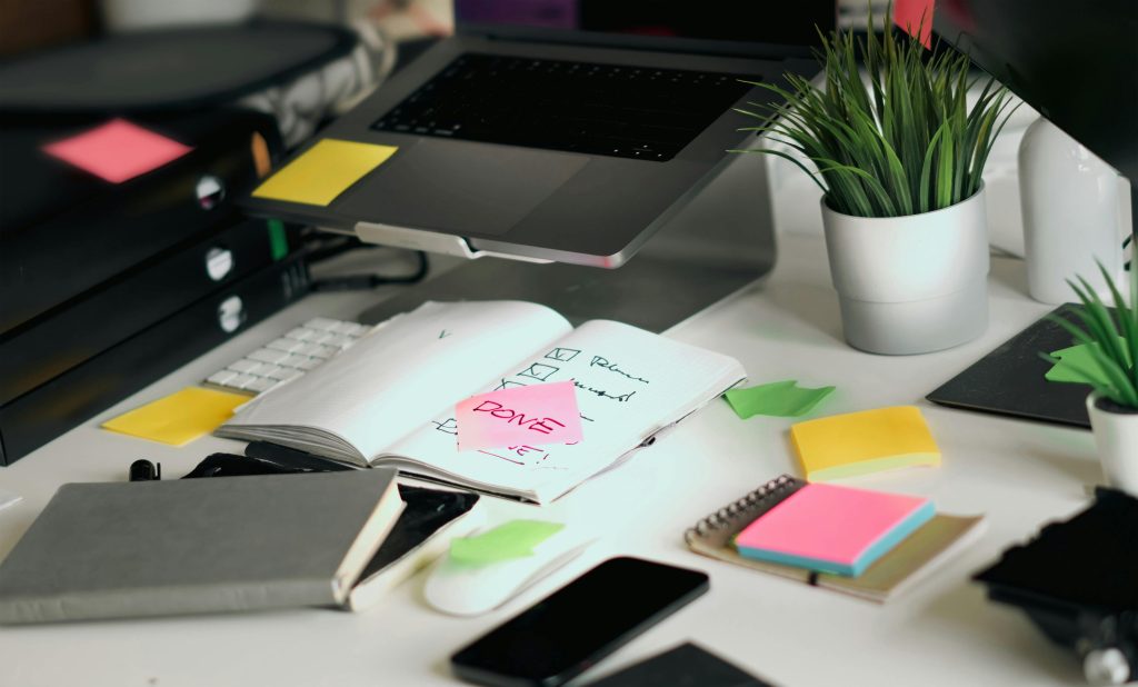 “Laptop on a stand, with a notebook featuring a pink ‘done’ sticky note, alongside a phone, additional sticky notes, and closed notebooks on a desk.