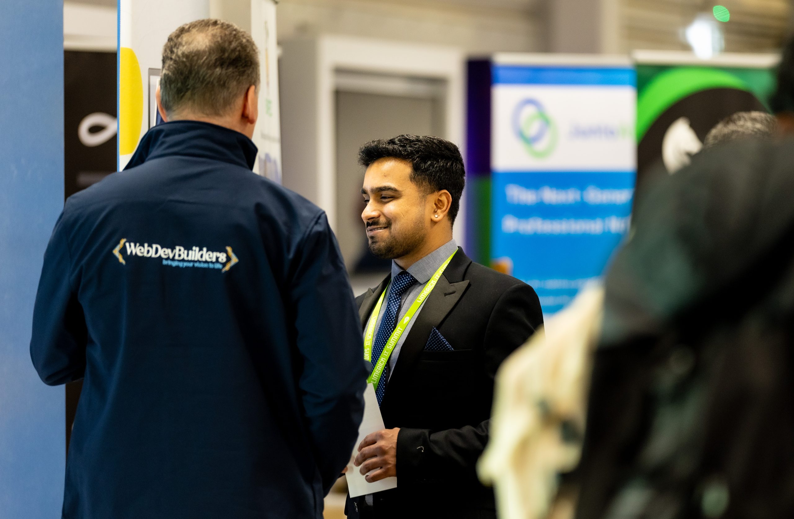 Back view of a WebDevBuilder staff member wearing a branded jacket, speaking with another attendee at the Limerick IT Summit.